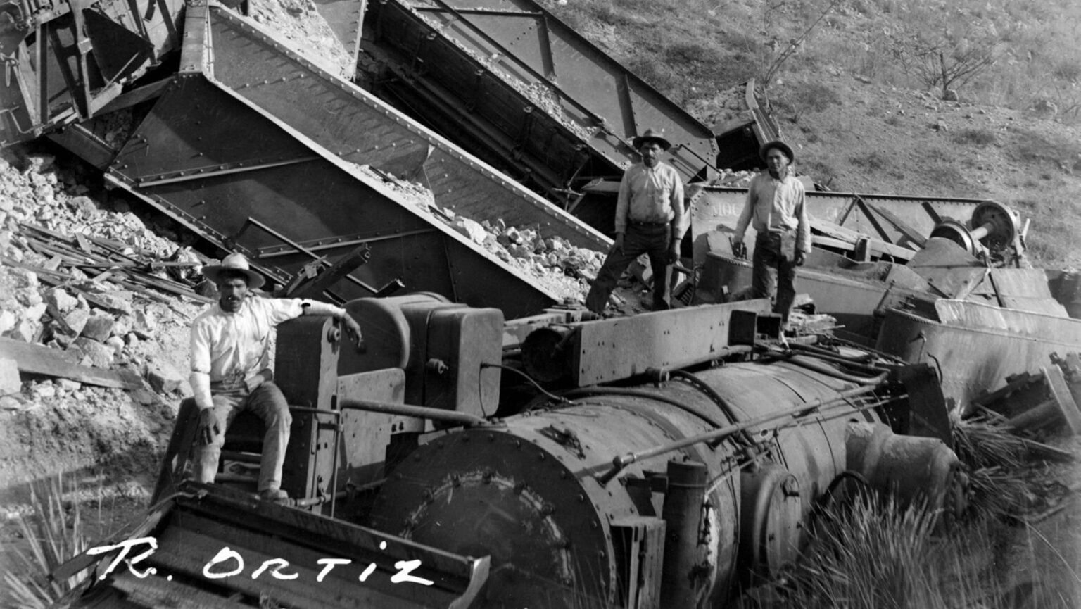 Fotografía histórica en blanco y negro de un descarrilamiento ocurrido en 1916; muestra una locomotora volcada y varios vagones dañados en una zona montañosa, con trabajadores de la época inspeccionando los restos sobre y alrededor del tren.
