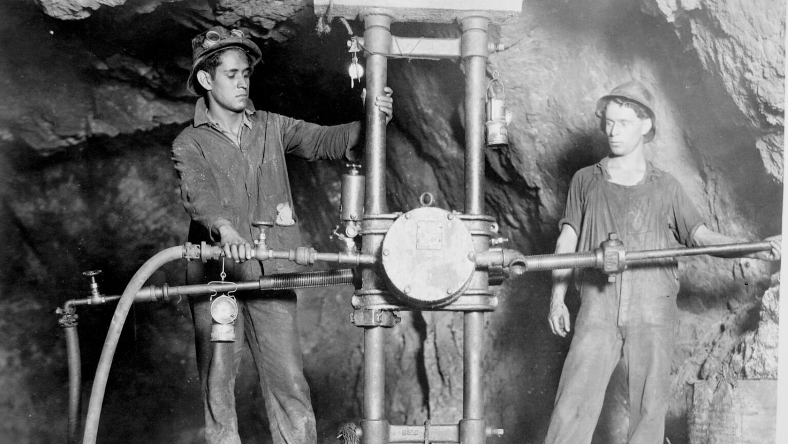 two young miners posing inside the Pilares mine in Nacozari, Sonora, on either side of a large drilling machine mounted on two vertical steel columns. The man on the left wears a cap with goggles on the front, a work shirt, and grips one of the columns and the air pipe. The man on the right wears a hard hat and overalls and holds a long horizontal rod from the machine. A thick air hose and metal pipes run along the bottom, carbide lamps hang from the equipment, and rough rock walls surround them.