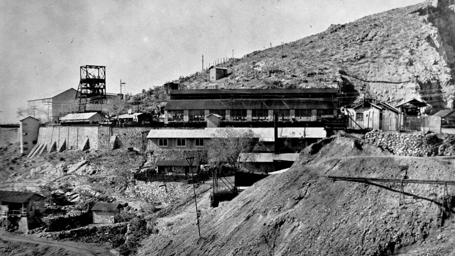 Historic mining complex in Pilares de Nacozari, Sonora, built into a rocky hillside, featuring several industrial structures including a tall metal headframe and long processing buildings with corrugated roofs; the uneven terrain is supported by stone retaining walls that reinforce the different levels of the facility.