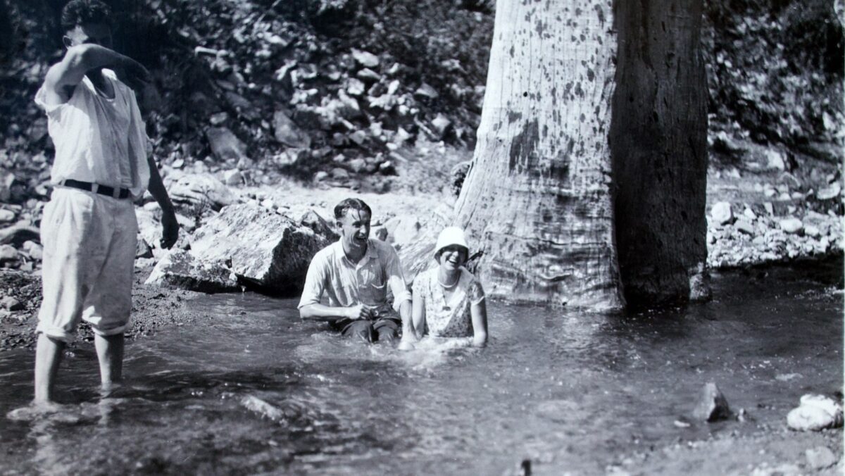 Black-and-white photograph from 1928 showing American tourists bathing in a shallow stream in Nacozari, Sonora, Mexico, beside the massive trunks of palm trees. One man stands in the water while a seated man and woman, wearing early 20th-century clothing and a sun hat, smile for the camera.