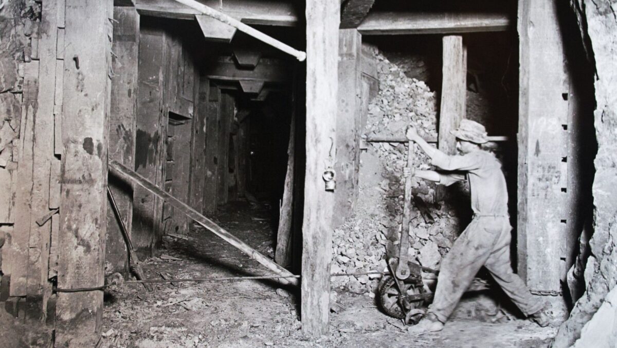 a miner working inside a timber-framed tunnel at the Pilares mine in Nacozari, Sonora. On the right, a man in a wide-brimmed hat and work clothes stands with his legs apart, gripping the long handle of a wheeled ore cart tipped toward a pile of broken rock. Heavy wooden posts and beams support the passage, a lamp hangs from one post, and a pipe runs overhead. To the left, a narrow timber-lined corridor recedes into the dark.
